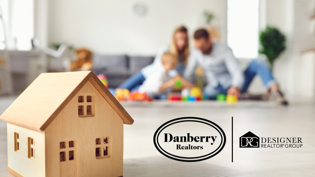 A small wooden toy house in the foreground with a blurred family playing in their spacious living room, featuring the Danberry Realtors and Designer Realtor Group logos.
