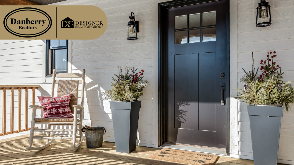 A welcoming front porch of a white farmhouse style home featuring a black front door a wooden rocking chair decorative holiday planters and the Danberry Realtors and Designer Realtor Group logos.